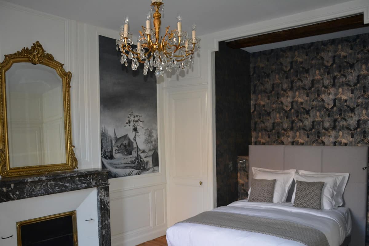 A bedroom with a bed, a chandelier, a fireplace, a large mirror, and a patterned wall.