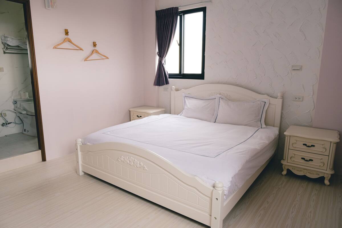 Un dormitorio con cama blanca, dos perchas de madera en la pared, una ventana con cortina y una mesita de noche con cajones.
