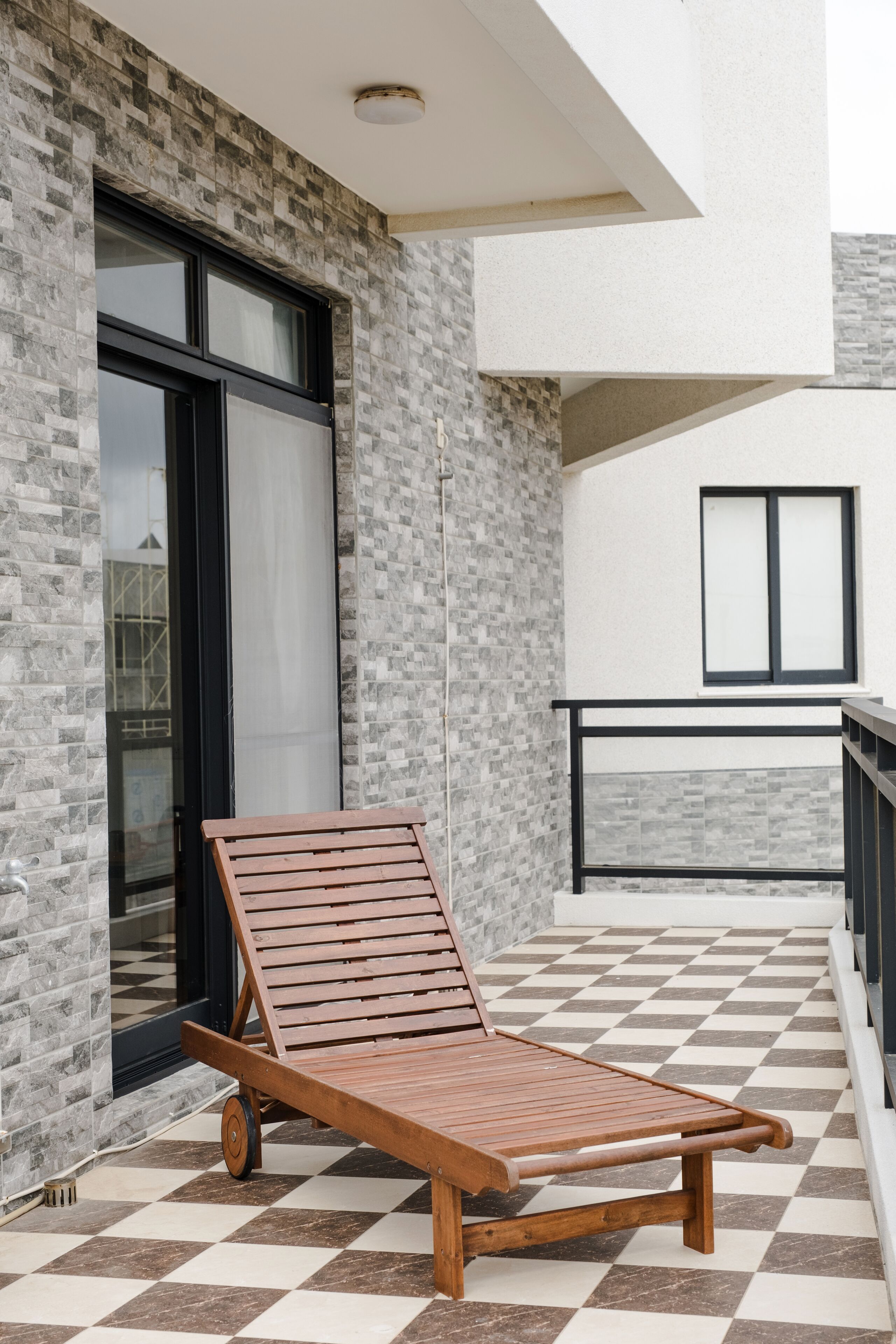 A wooden lounge chair on a checkered floor, a balcony with a black railing, and a building facade with a mix of brick and smooth surfaces.