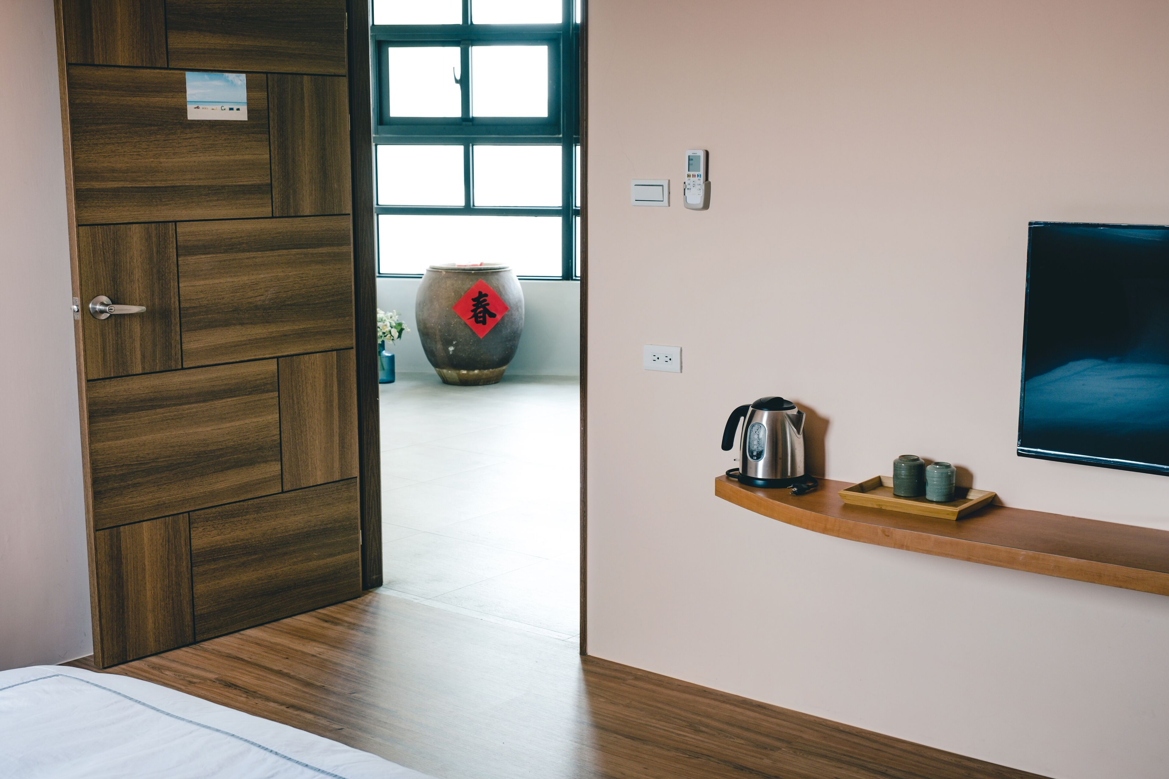 A modern hotel room with a wooden door, a large window, a wall-mounted TV, and a shelf with a kettle and cups.
