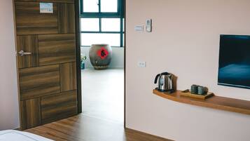 A modern hotel room with a wooden door, a large window, a wall-mounted TV, and a shelf with a kettle and cups.