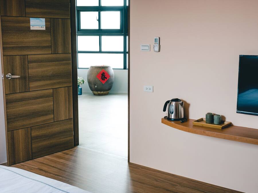 A modern hotel room with a wooden door, a large window, a wall-mounted TV, and a shelf with a kettle and cups.