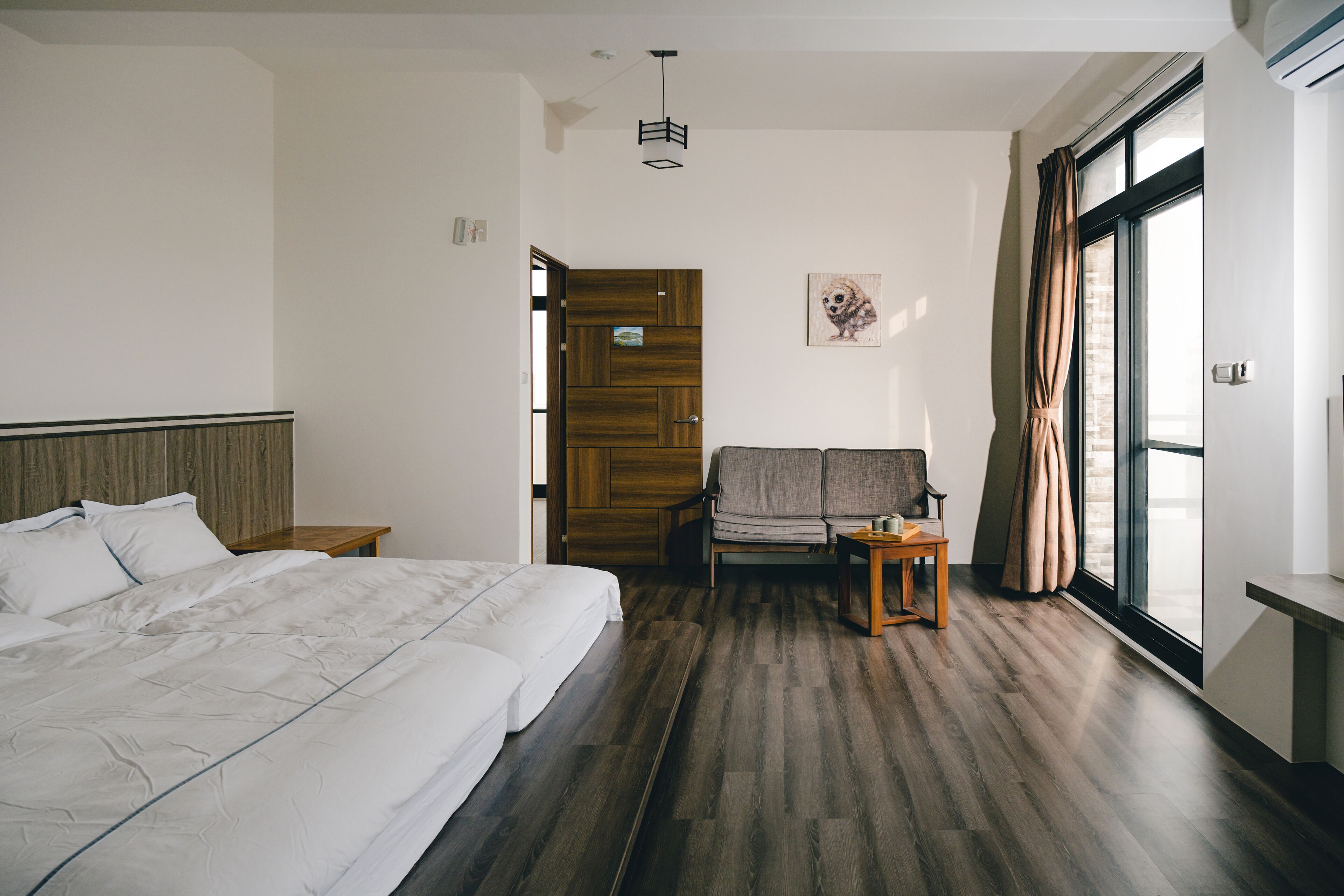 A hotel room with a large bed, a wooden floor, a sofa, a small table, and a window with curtains.