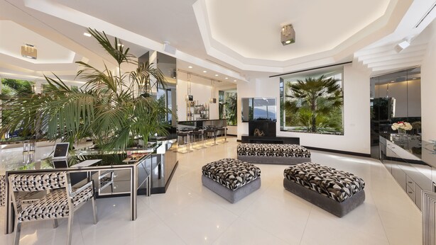 A modern, spacious living area with a high ceiling, a central seating area with patterned cushions, a bar area, and a large window offering a view of greenery. - Villa Silvana Relais (Sorrento)