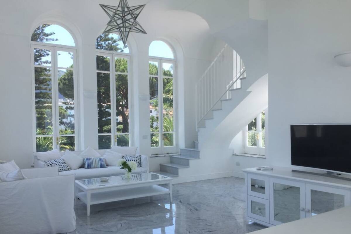 A spacious living room with a white sofa, a coffee table, and a TV on a white cabinet. There are large windows with a star-shaped decoration hanging from the ceiling.