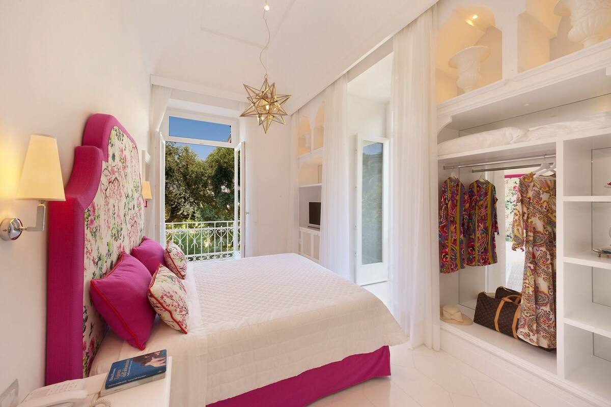 A bedroom with a bed, a nightstand, a lamp, a wardrobe, and a window with a view of trees.