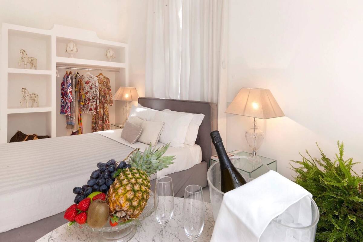 A hotel room with a bed, a bedside table with a bottle of champagne, a fruit bowl, and a lamp.