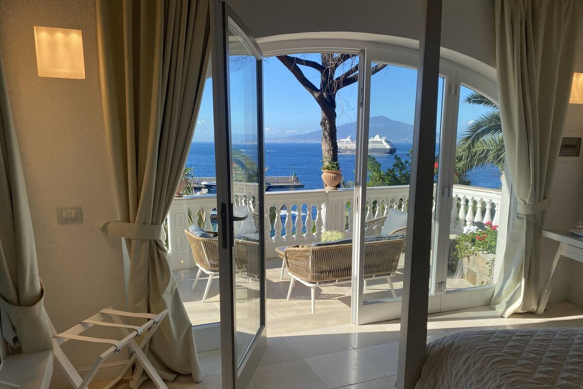 A balcony with wicker furniture, a potted plant, and a view of the sea and cruise ships.