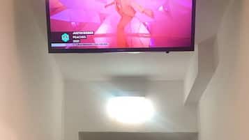 A hallway with a flat-screen TV mounted on the wall displaying a music video. A white door is visible at the end of the hallway.