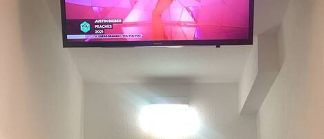 A hallway with a flat-screen TV mounted on the wall displaying a music video. A white door is visible at the end of the hallway.