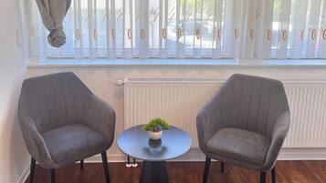 A room with two grey armchairs, a small round table with a plant, and a window with patterned curtains.