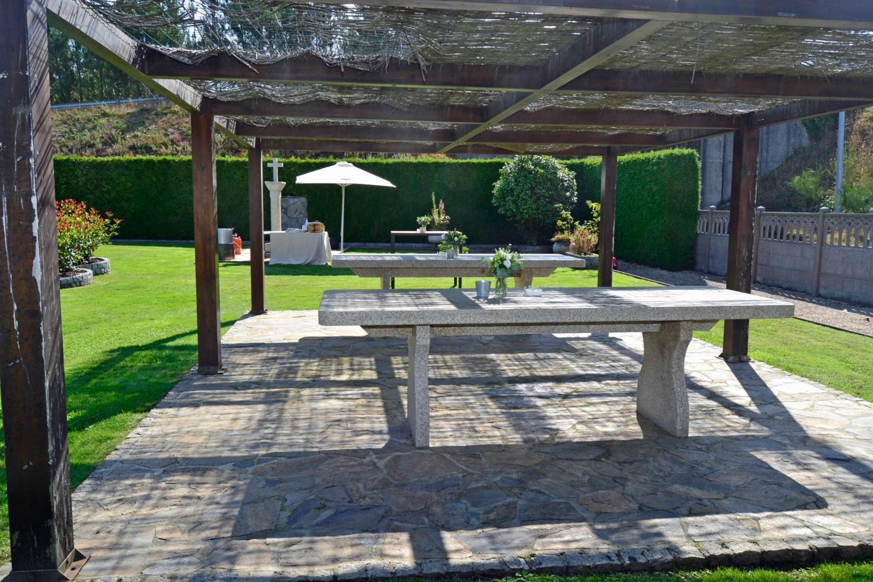 An outdoor patio area with a stone table and benches, surrounded by a hedge and a wooden pergola.