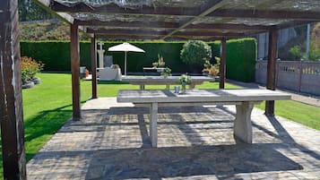 An outdoor patio area with a stone table and benches, surrounded by a hedge and a wooden pergola.