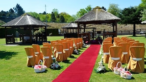Une cérémonie de mariage en plein air, avec un tapis rouge, des chaises orange et deux pergolas à toit de chaume.