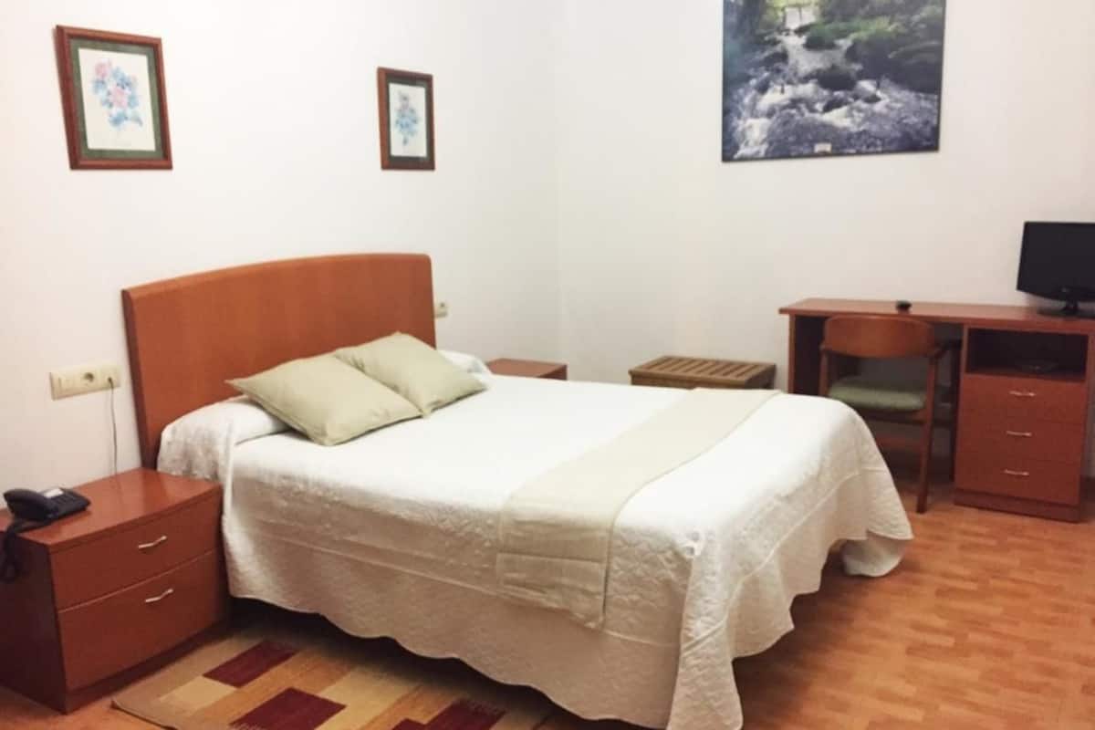 Un dormitorio ordenado con cama, mesitas de noche, escritorio y televisor.