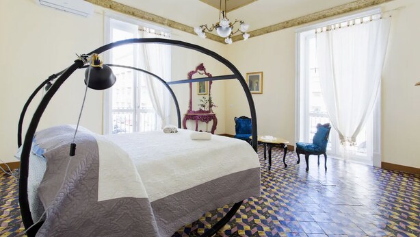A room with a bed under a circular canopy, a chandelier, a blue chair, and a patterned floor. - Artisan Maison (Palermo)