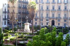A plaza with a statue, palm trees, and a building with balconies. - Artisan Maison (Palermo)