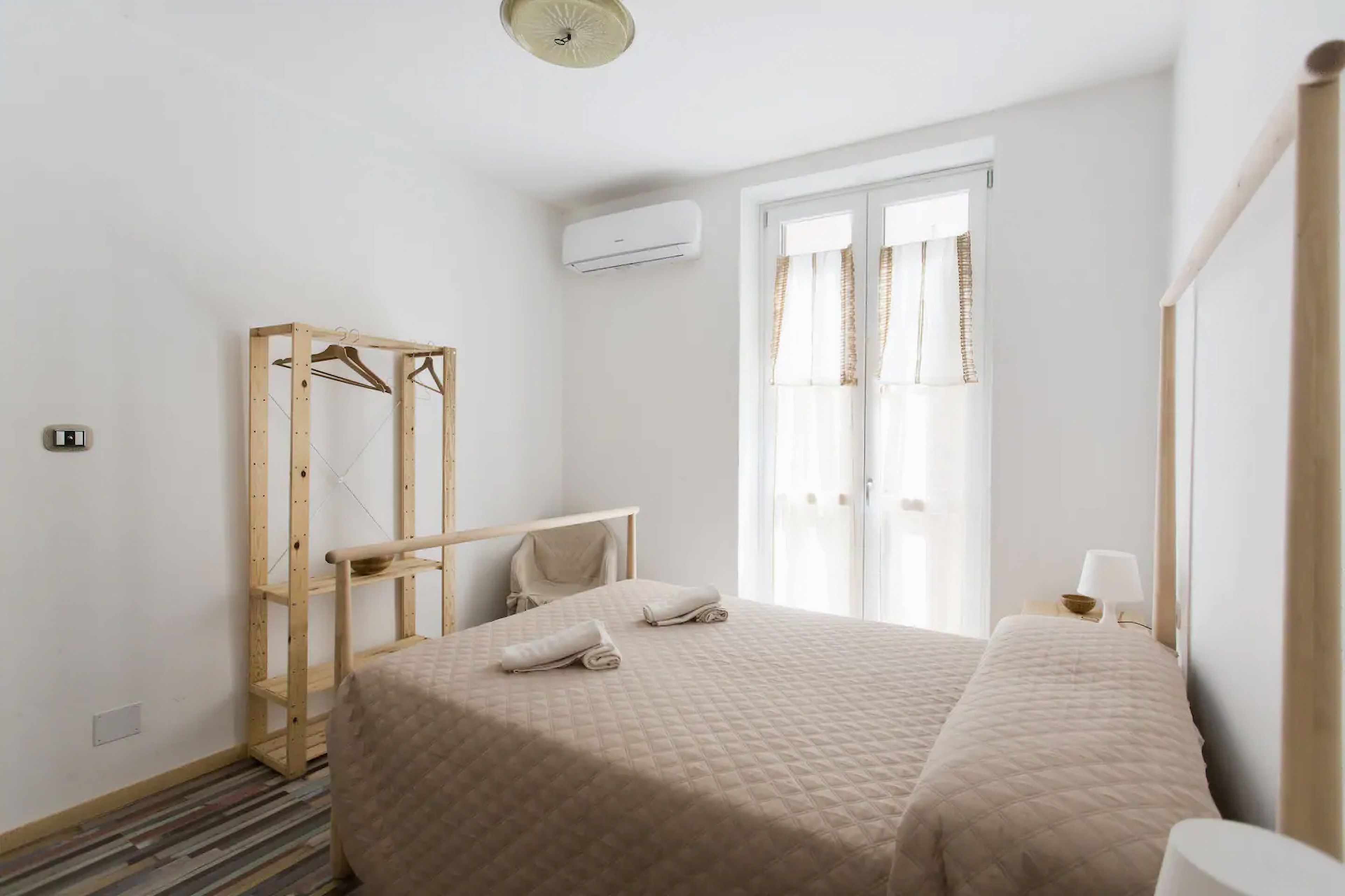 A bedroom with two beds, a wooden wardrobe, a bedside table with a lamp, and a window with curtains.