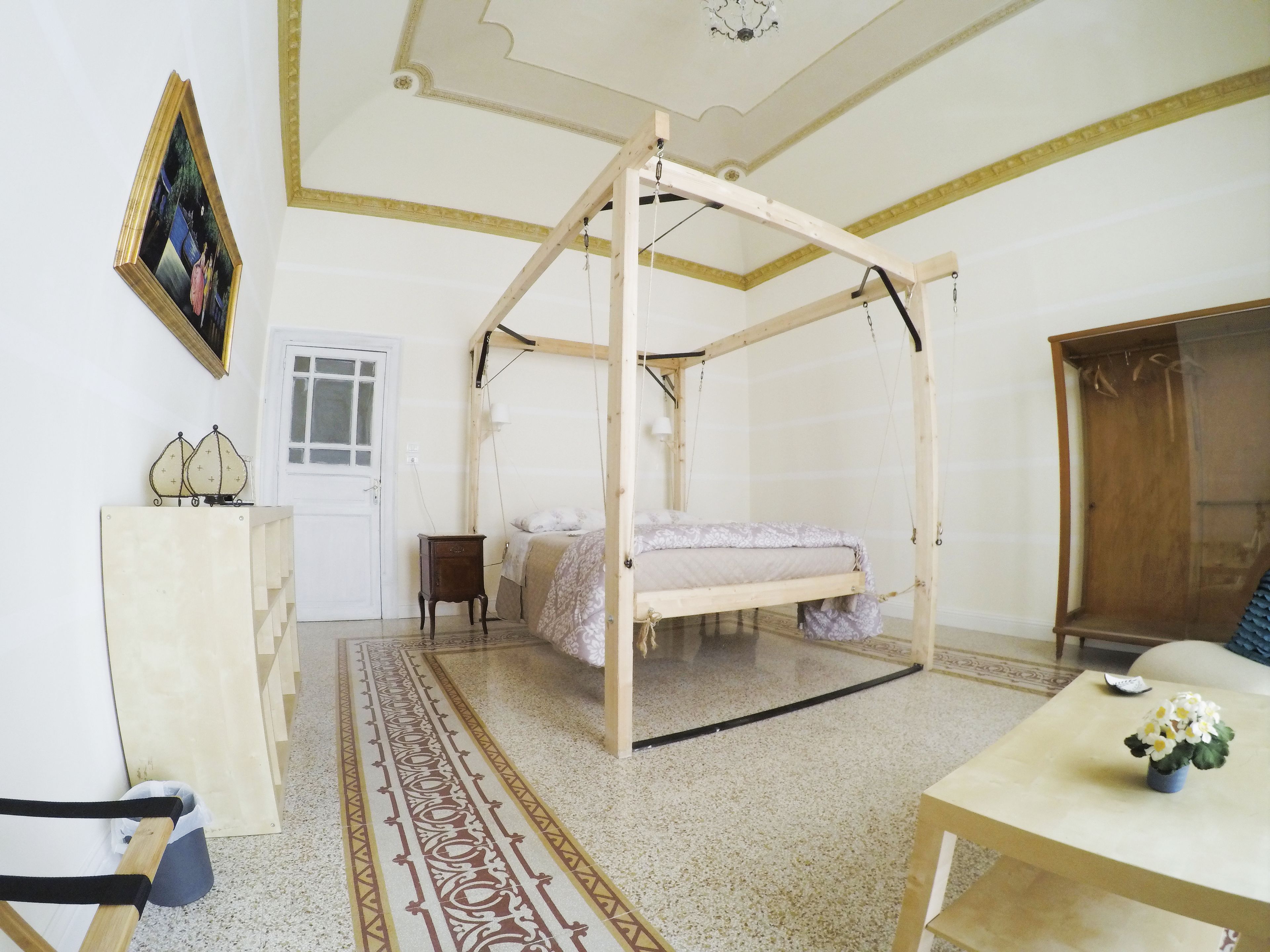 Ein Zimmer mit einem hölzernen Baldachinbett, einem kleinen Tisch mit Blumen, einem Stuhl, einem Schreibtisch und einem gerahmten Bild an der Wand.