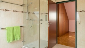 Un baño moderno con ducha envuelta en vidrio, un perchero con una toalla verde y una puerta de madera que da a otra habitación.