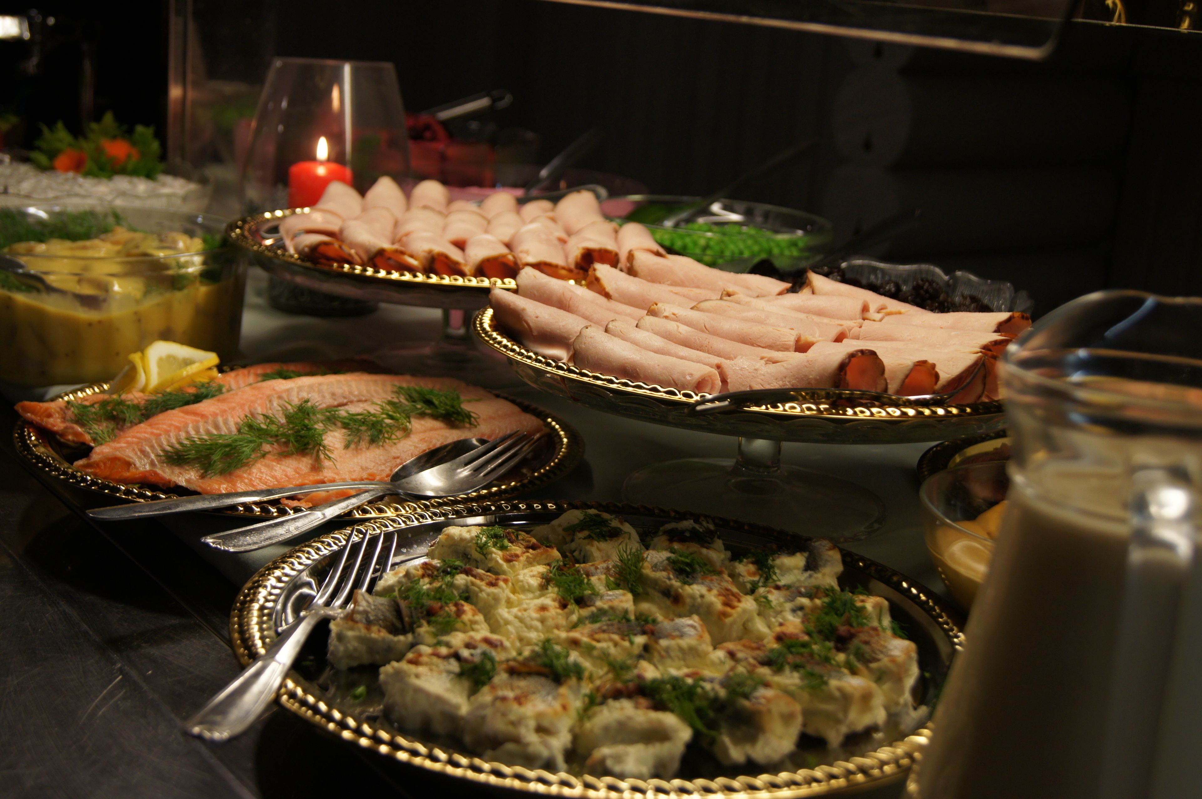 A buffet table with various dishes, including salmon, rolls, and a casserole, with serving utensils and a candle in the background.