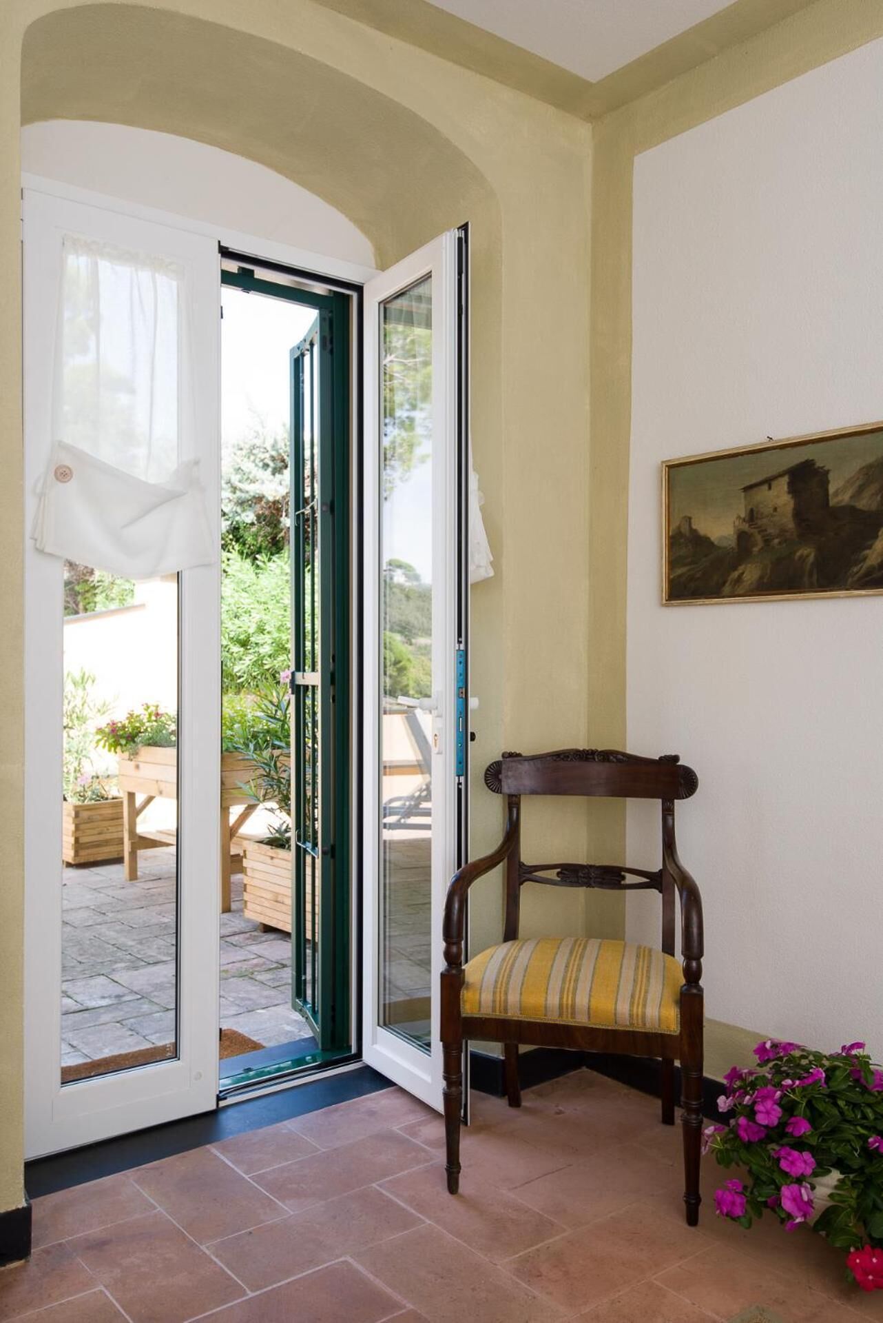 A room with a wooden chair, a painting on the wall, and a view through a glass door to an outdoor area with plants and a bench.