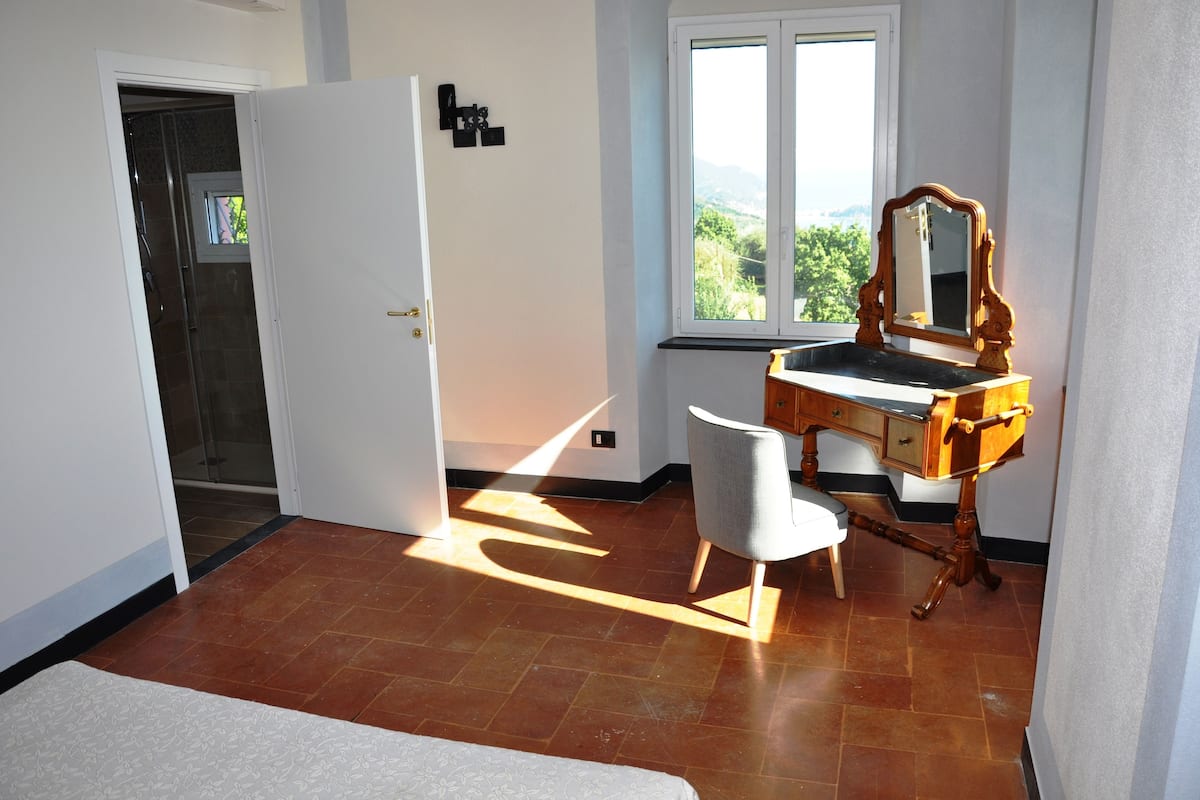 A room with a wooden vanity, a chair, a bed, a window with a view, and a door leading to a shower.