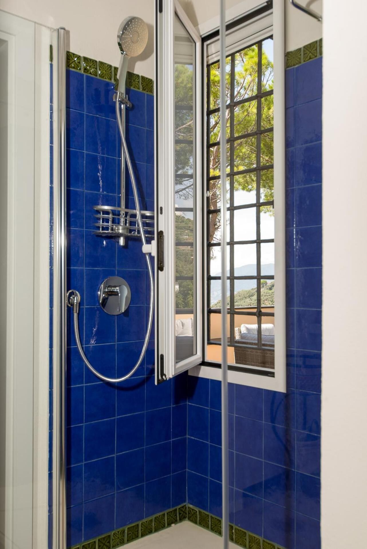 A shower with a glass door, blue tiles, and a view of trees and mountains through the window.