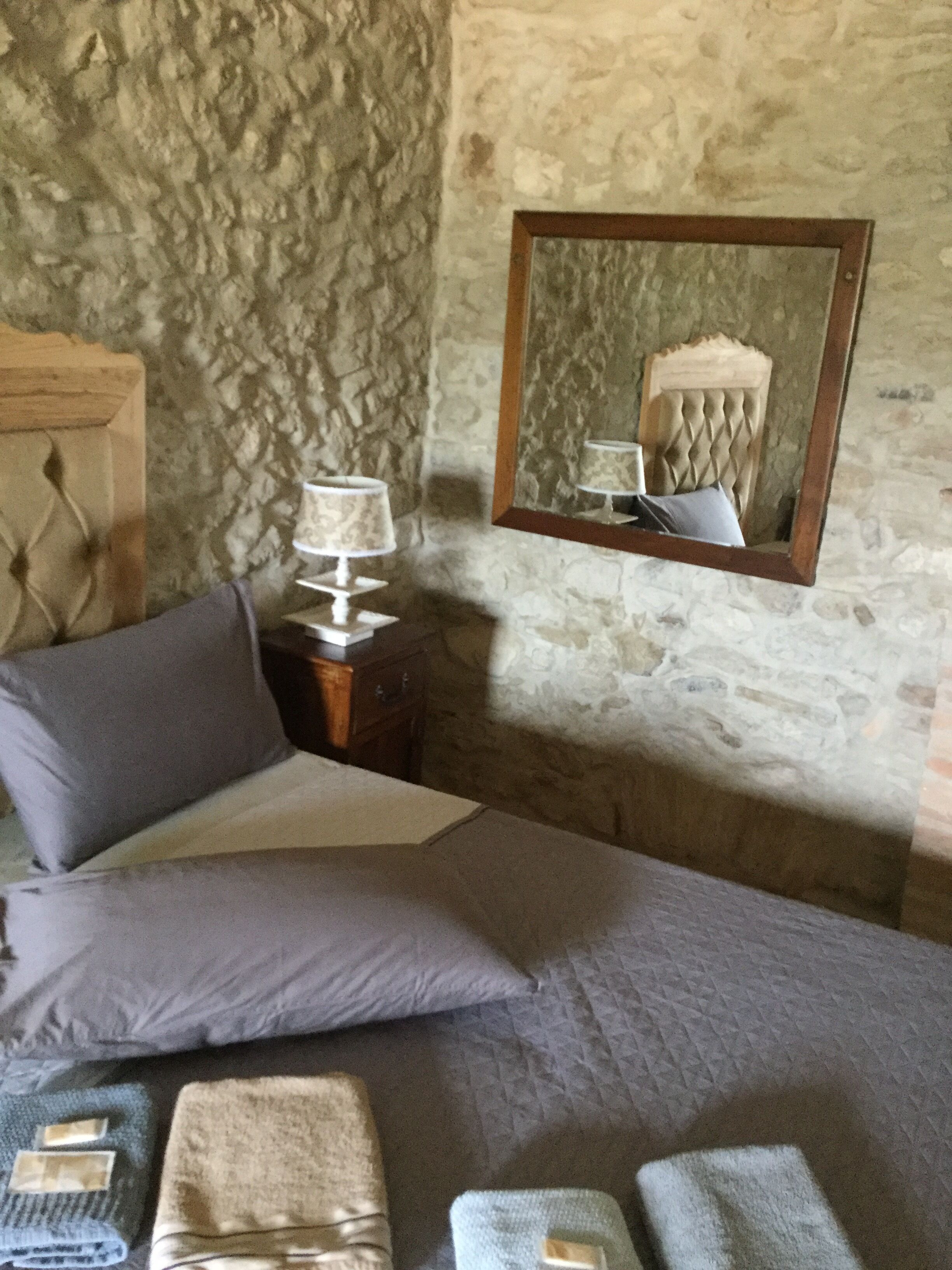 Une chambre avec un mur en pierre, une tête de lit en bois, un lit avec une literie grise, une table de chevet avec une lampe et un miroir sur le mur.