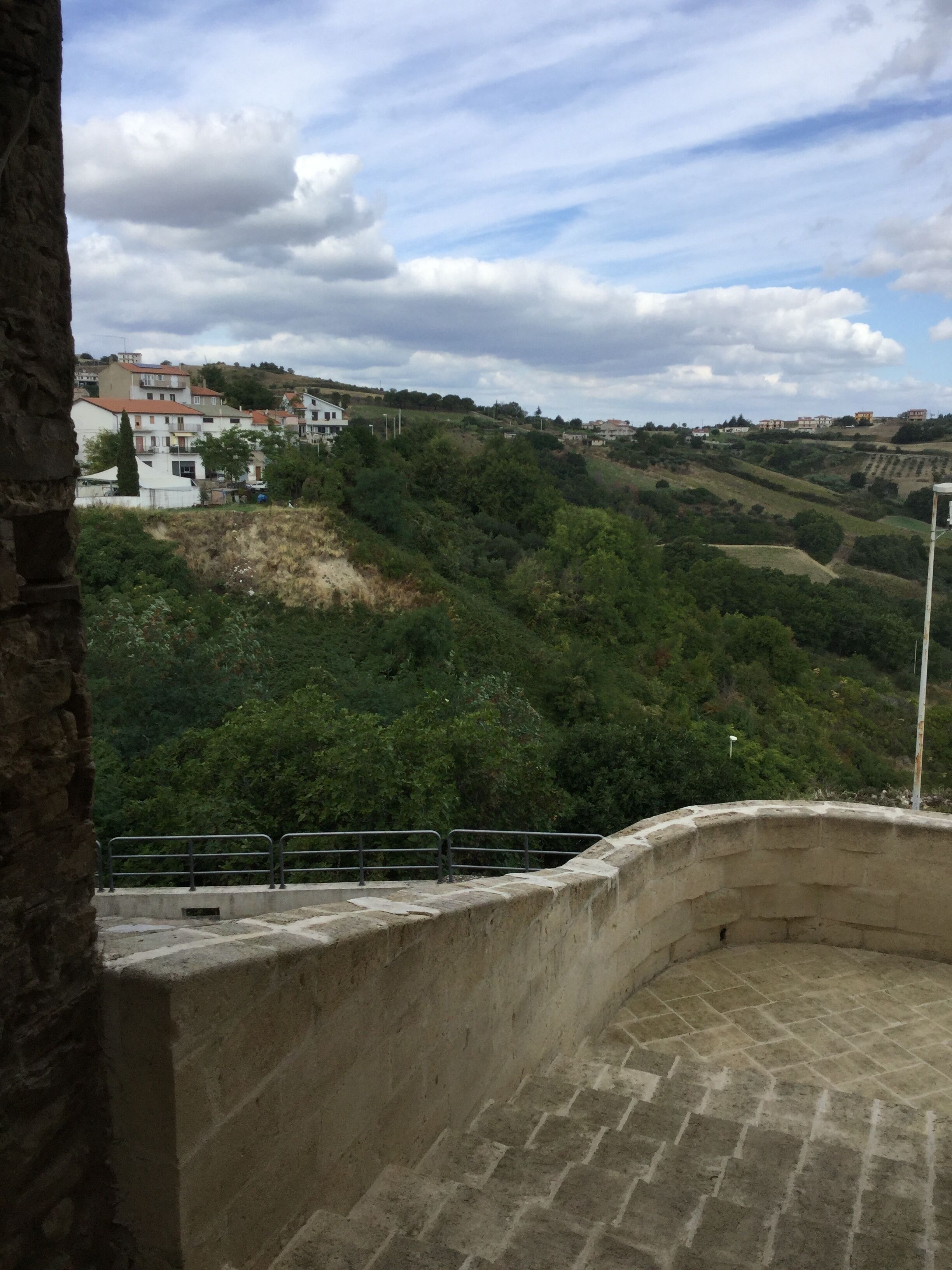 Una construcció de pedra amb un mur corbat i un paviment, amb vista a un paisatge amb turons, edificis i vegetació.