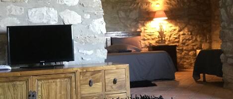 A room with a stone wall, a television on a wooden cabinet, a bed with a lamp, and a green carpet.