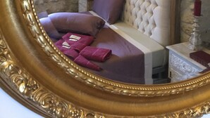 A bedroom with a stone wall, a bed with a tufted headboard, a nightstand, a lamp, and a mirror with a gold frame.