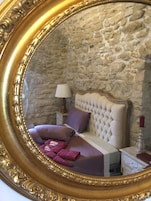 A bedroom with a stone wall, a bed with a tufted headboard, a nightstand, a lamp, and a mirror with a gold frame.
