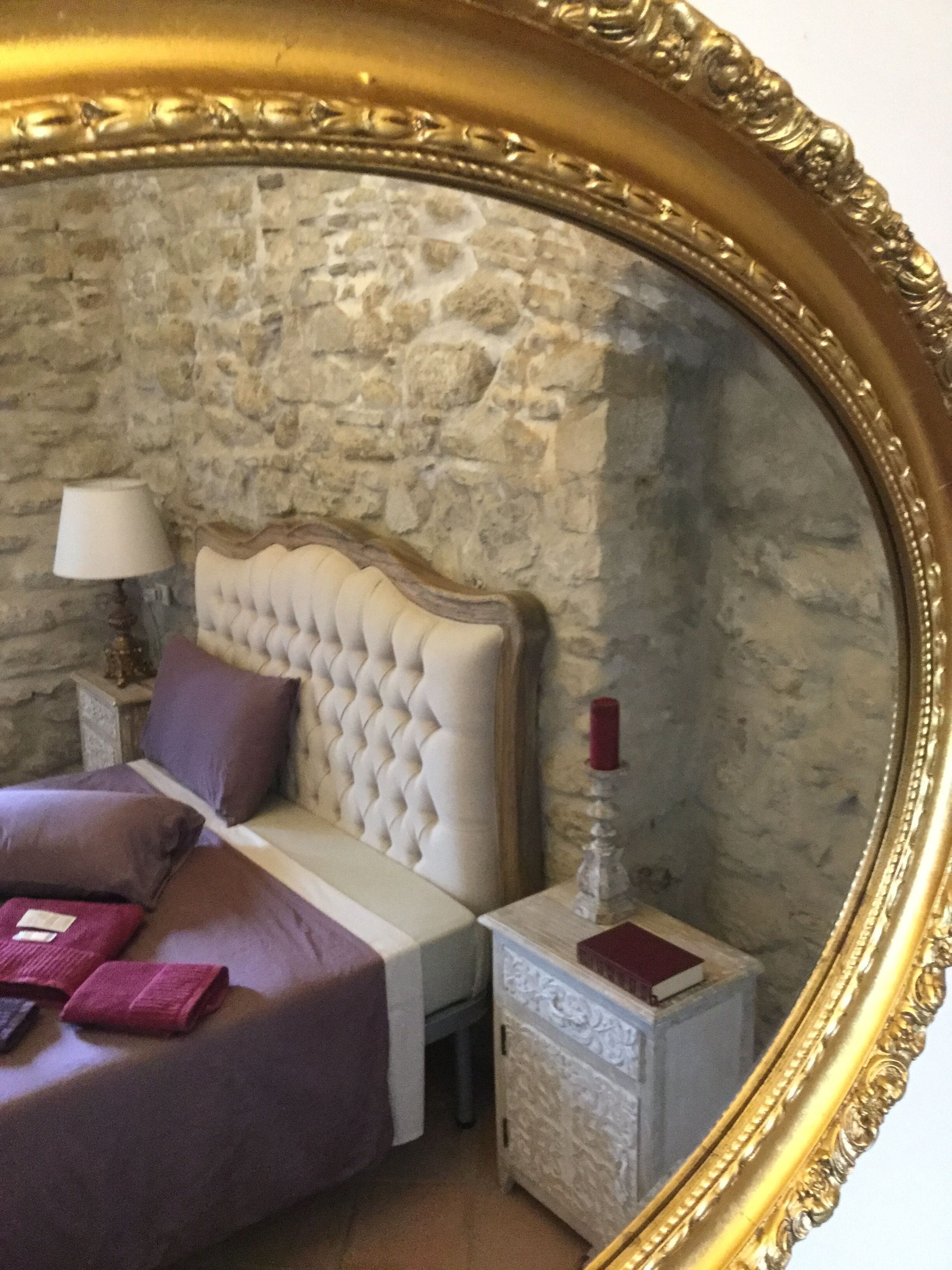 A bedroom with a stone wall, a tufted headboard, a bed with purple bedding, a nightstand with a lamp, and a red candle.