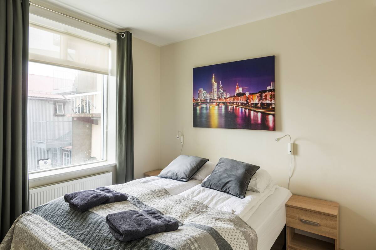 Ein ordentlich bezogenes Bett mit einer gemusterten Tagesdecke, zwei Kissen und gefalteten Handtüchern. Über dem Bett hängt ein Stadtbild.