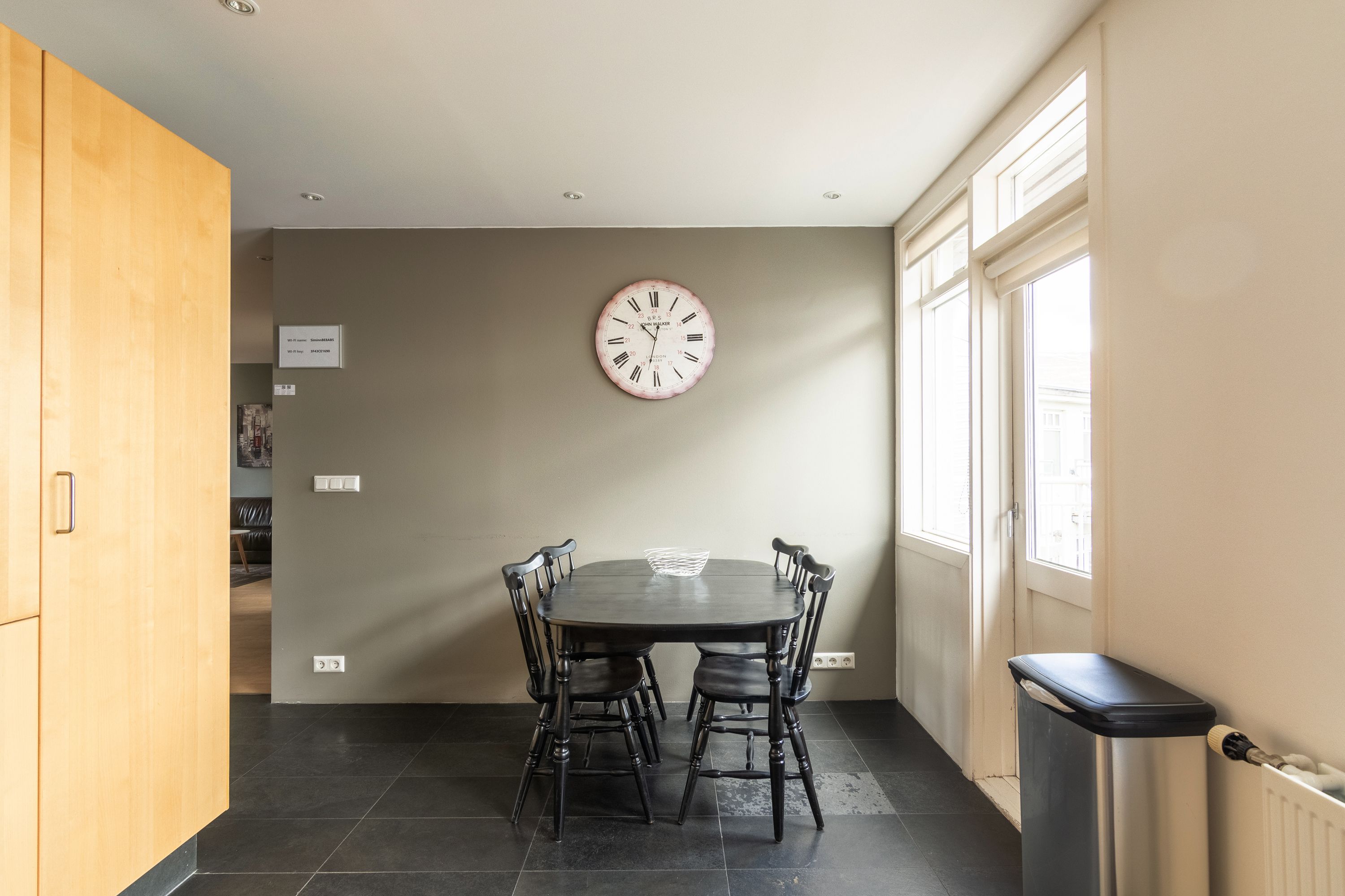 Un comedor con una mesa redonda y sillas, un reloj de pared y un cubo de basura.