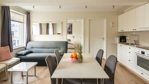 A modern living room with a grey sofa, a white table with a fruit bowl, and a kitchen area with white cabinets and a stove.