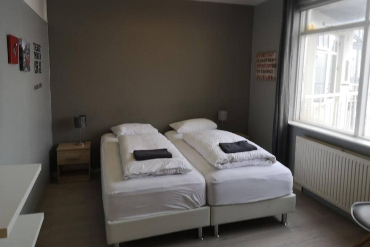 Ein modernes Hotelzimmer mit zwei Einzelbetten, einem Nachttisch mit Lampe, einem kleinen Schreibtisch und einem Fenster mit Blick auf Gebäude.