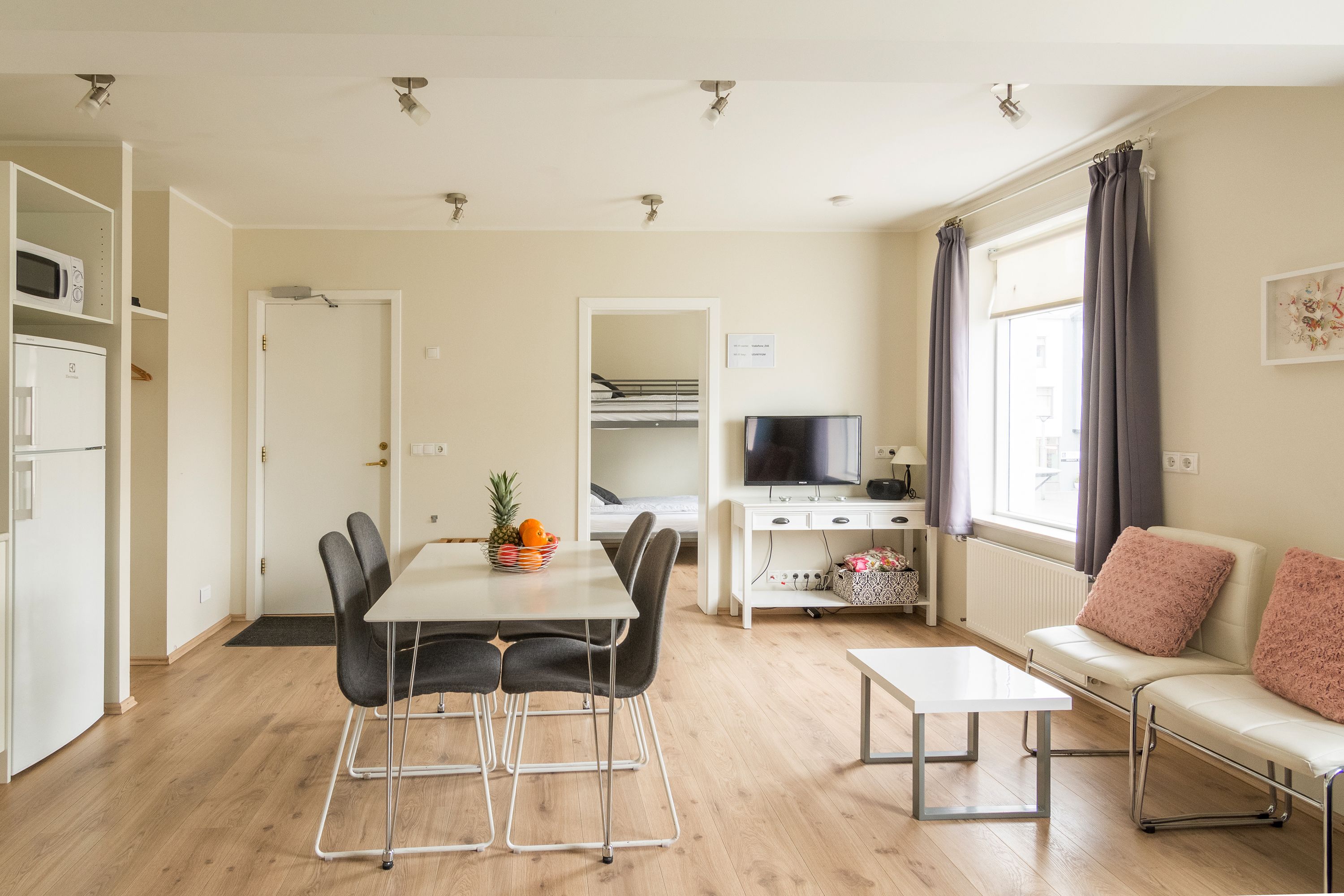 A modern, well-lit room with a dining table, chairs, a sofa, a TV, and a kitchen area with a microwave.