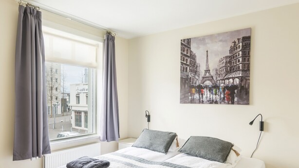 A hotel room with a bed, grey curtains, a window with a view of buildings, and a painting of Parisian architecture.