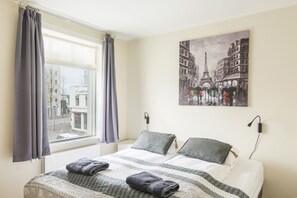 Habitación de hotel con cama, cortinas grises, ventana con vistas a edificios y un cuadro de arquitectura parisina.