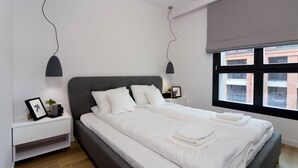 Un dormitorio moderno con una cama grande, una mesita de noche, una ventana con persianas y vistas a edificios.