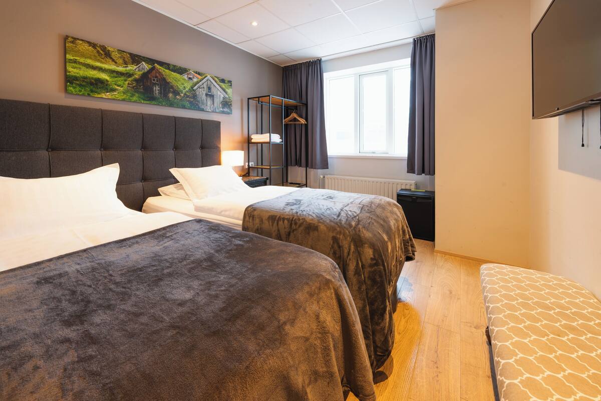A hotel room with two beds, a large window with curtains, a television mounted on the wall, and a painting above the bed.