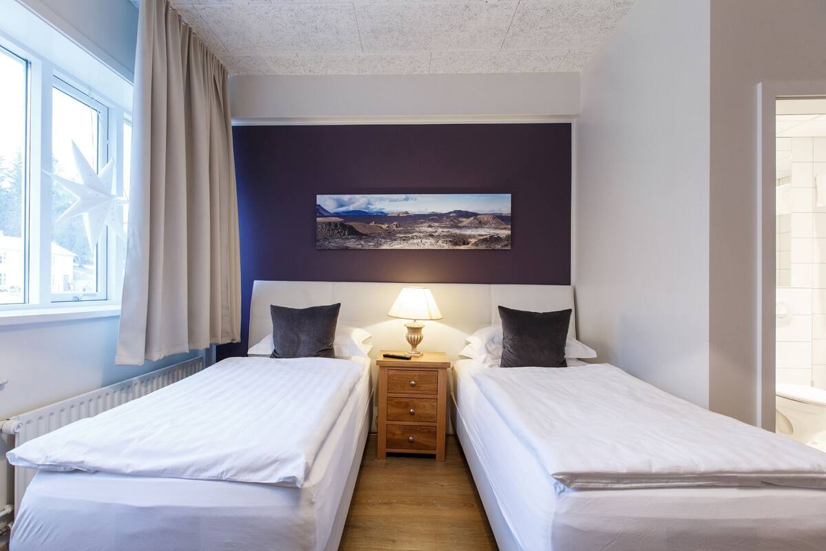 A hotel room with two beds, a nightstand with a lamp, a painting on the wall, and a window with curtains.
