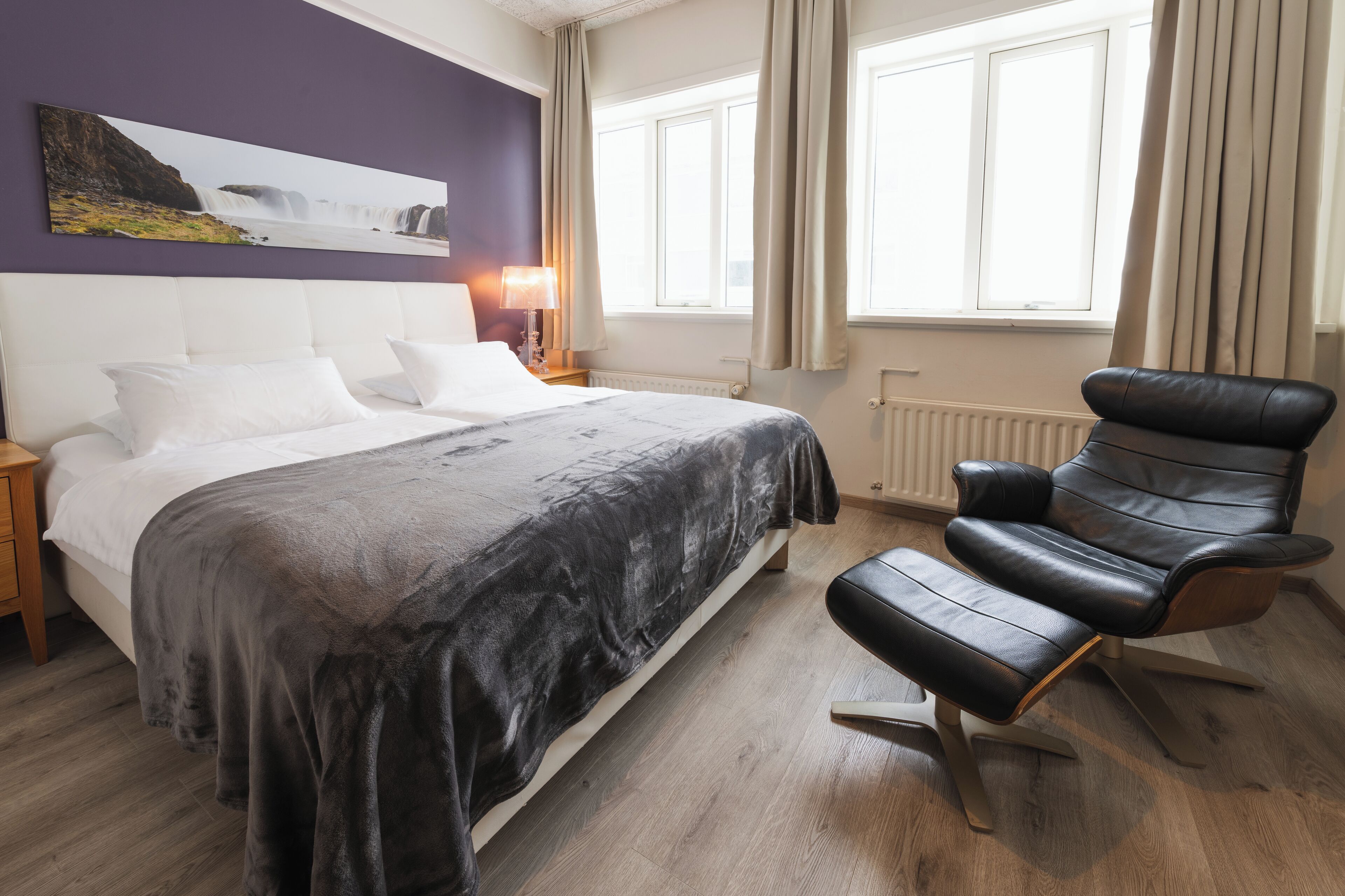 Ein Schlafzimmer mit Bett, Sessel, Nachttisch, Lampe und einem Fenster mit Vorhängen.