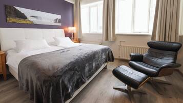 Ein Schlafzimmer mit Bett, Sessel, Nachttisch, Lampe und einem Fenster mit Vorhängen.