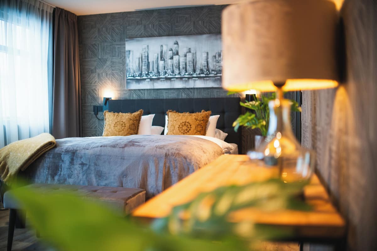 A hotel room with a bed, a bedside table with a lamp, a cityscape artwork on the wall, and a window with curtains.