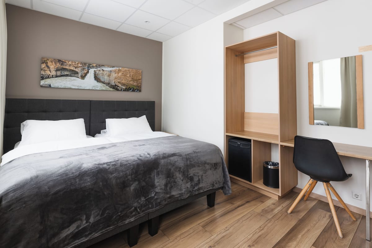 A modern hotel room with a large bed, a desk with a chair, a wooden wardrobe, and a painting on the wall.