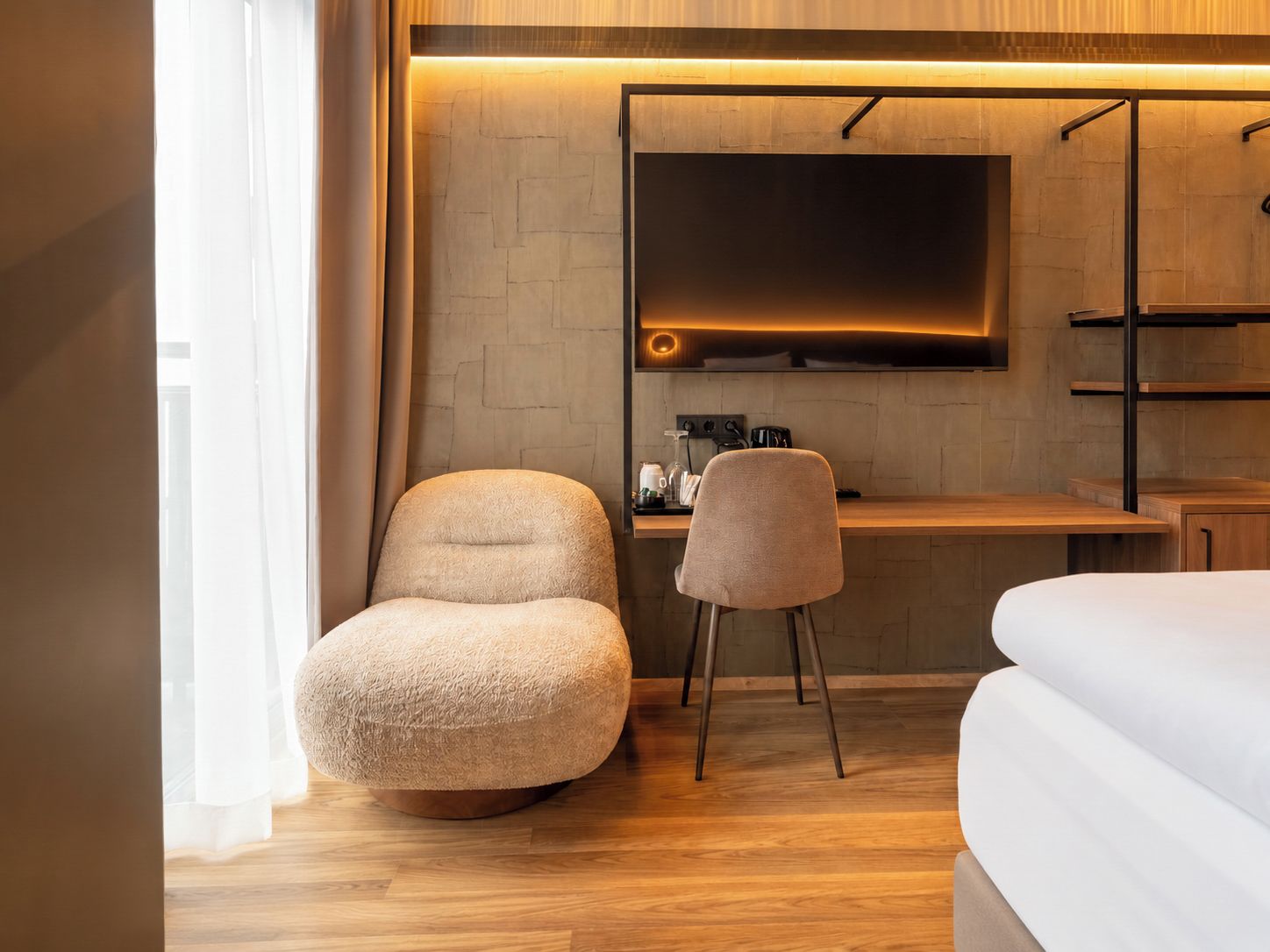 A modern hotel room with a wooden floor, a large bed, a sofa, a desk with a flat-screen TV, and a coffee machine.
