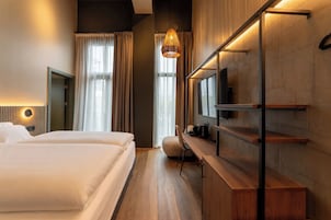 A modern hotel room with a large bed, a desk with a chair, a TV mounted on the wall, and a hanging light fixture.